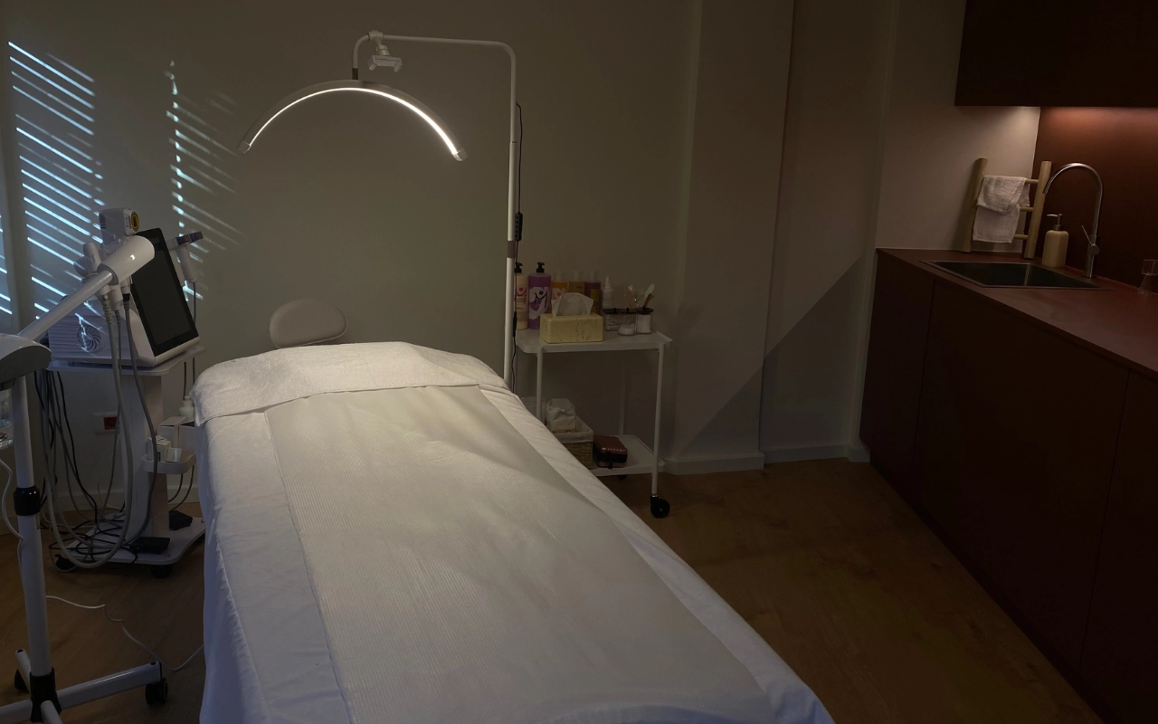 Ένα εστιασμένο δωμάτιο θεραπείας με λευκό κρεβάτι κάτω από τοξωτό LED φωτιστικό, επαγγελματικό εξοπλισμό αισθητικής με ψηφιακή οθόνη, προϊόντα skincare σε τροχήλατο, και σκούρα ξύλινα ντουλάπια με νεροχύτη στο βάθος.