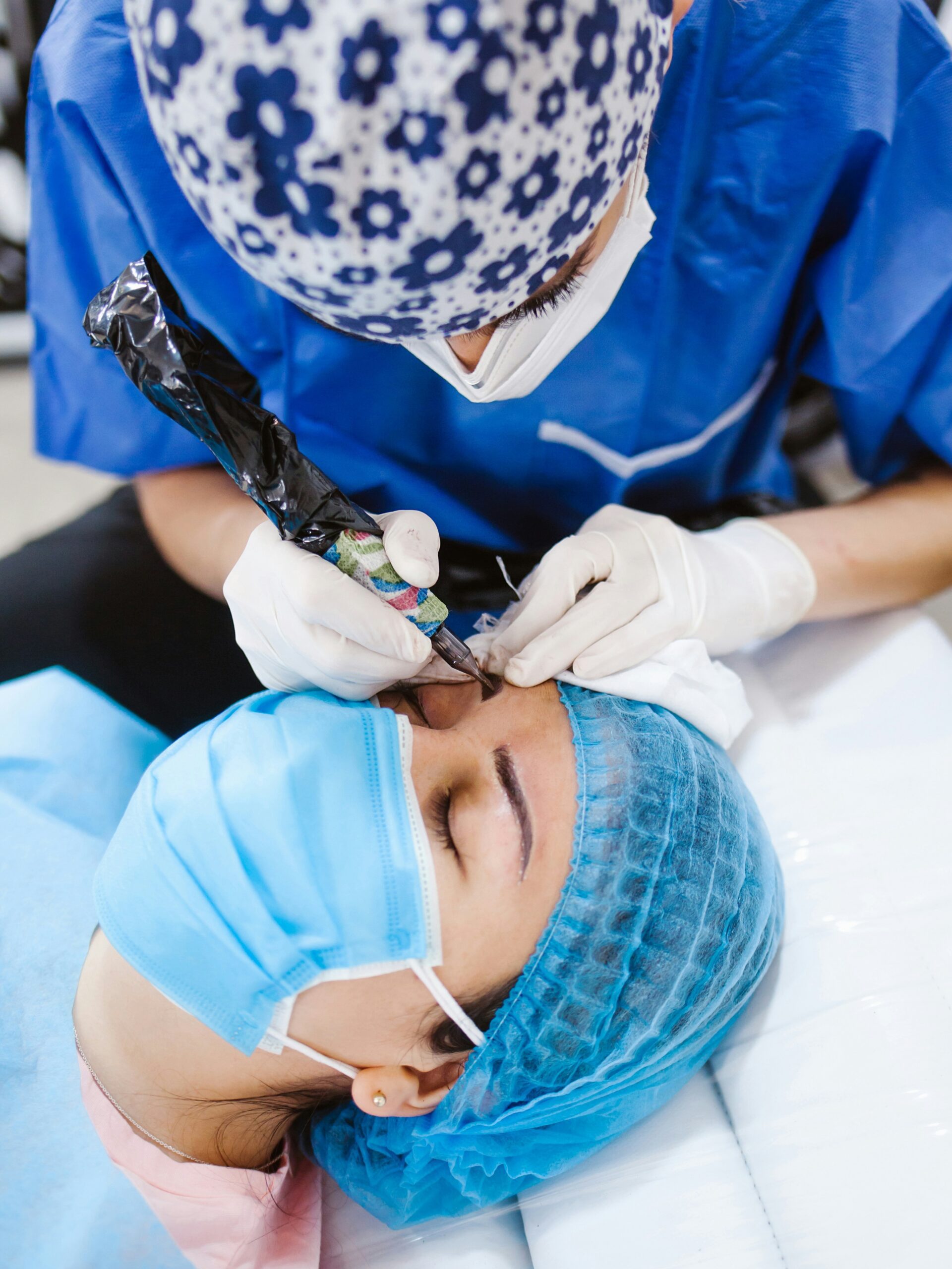 Κοντινό πλάνο μιας γυναίκας που κάνει microblading, ξαπλωμένη σε κρεβάτι θεραπείας φορώντας μάσκα, ενώ ο τεχνίτης εργάζεται στα φρύδια της