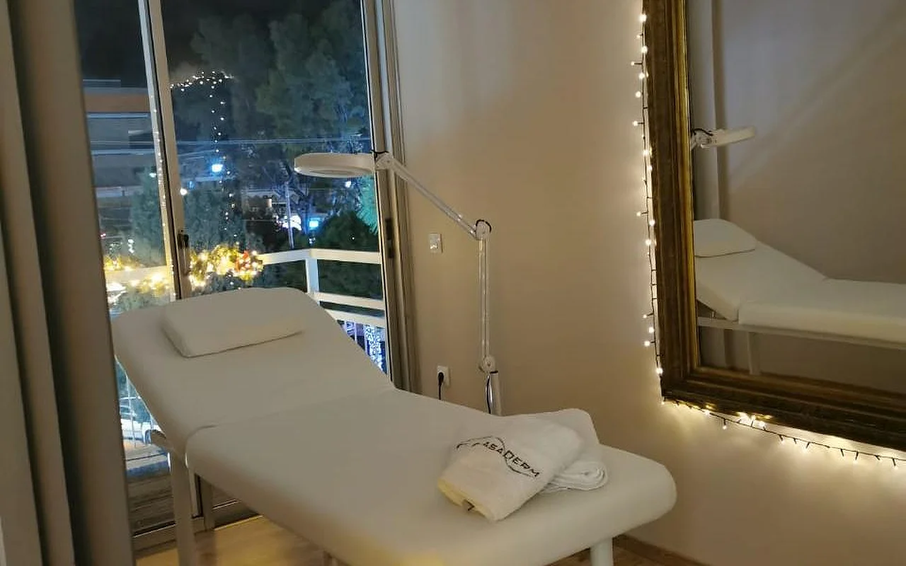 Ένα απαλά φωτισμένο δωμάτιο θεραπείας με λευκό κρεβάτι, πετσέτα με το λογότυπο CasaDerm, μεγεθυντικό φωτιστικό, φωτάκια που πλαισιώνουν χρυσό καθρέφτη, και μπαλκονόπορτα με θέα στο βραδινό τοπίο της Γλυφάδας.