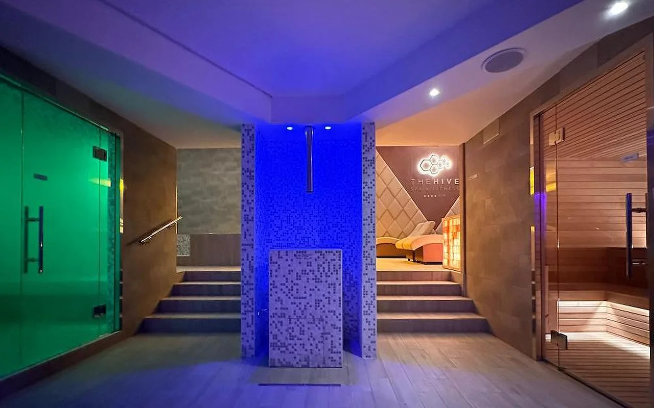 Η θερμική ζώνη ενός πολυτελούς spa με δραματικό μπλε και πράσινο ατμοσφαιρικό φωτισμό. Ο χώρος διαθέτει ξύλινη φινλανδική σάουνα, είσοδο ατμόλουτρου και ένα κεντρικό μωσαϊκό στοιχείο με σκαλοπάτια που οδηγούν σε ζώνες χαλάρωσης.
