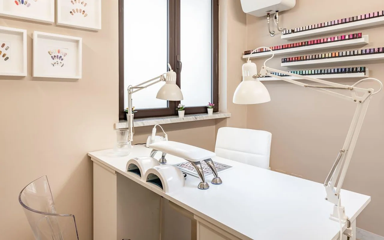 Ένας αποκλειστικός πάγκος νυχιών στο AG The Beauty Room's με καθαρό λευκό γραφείο, διπλές λάμπες UV, ρυθμιζόμενα φωτιστικά εργασίας και ράφια που εκθέτουν σειρές από χρωματιστά βερνίκια νυχιών. Κορνιζαρισμένα σχέδια nail art κρέμονται στους τοίχους σε ζεστούς τόνους.