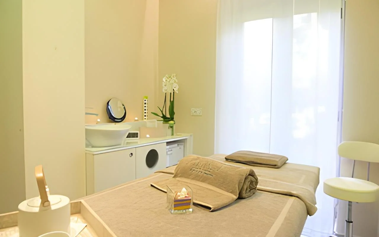 Ένας ήρεμος χώρος θεραπείας στο Alice Beauty Clinique με κρεβάτι ντυμένο σε απαλά taupe λευκά είδη, λευκές ορχιδέες στον πάγκο, κεριά δίπλα στον νιπτήρα και διάφανες κουρτίνες που φιλτράρουν το φυσικό φως μέσα από ένα μεγάλο παράθυρο.