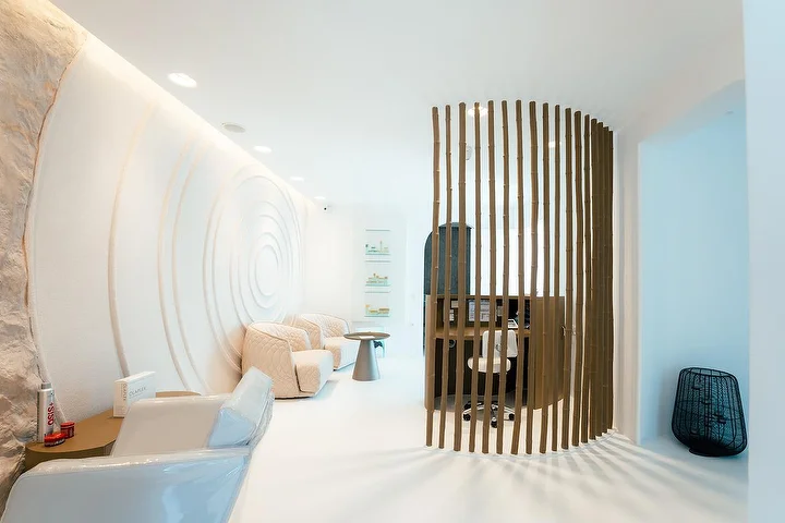 Ένας ήσυχος χώρος spa με αίσθηση σπηλιάς, με καμπυλωτούς λευκούς τοίχους, ο ένας σκαλισμένος σε ομόκεντρους κυματισμούς, με απαλό έμμεσο φωτισμό, αναπαυτικές πολυθρόνες σε χρώμα κρεμ και ένα καμπυλωτό ξύλινο διαχωριστικό από πηχάκια που οριοθετεί μια ήσυχη γωνιά θεραπείας.