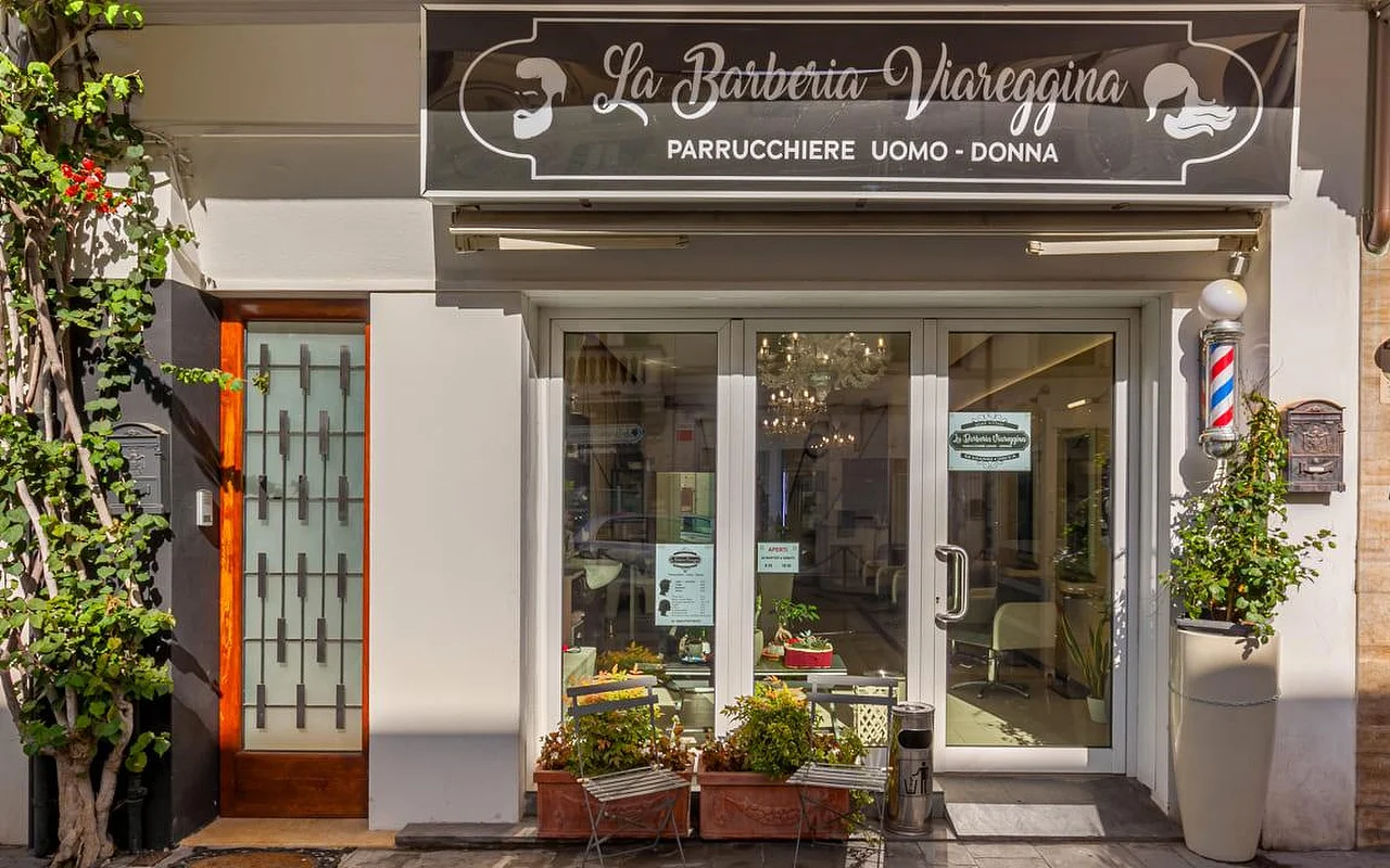 Η πρόσοψη του La Barberia Viareggina μια ηλιόλουστη μέρα, με μια κλασική κόκκινη, λευκή και μπλε κολόνα κουρείου δεξιά από τις γυάλινες πόρτες. Γλάστρες και αναρριχώμενη πρασινάδα πλαισιώνουν την είσοδο, ενώ μέσα διακρίνεται ένας πολυέλαιος μέσα από τα τζάμια.