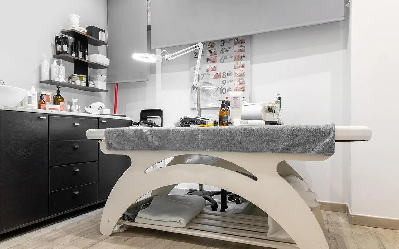 Μια συμπαγής καμπίνα αισθητικής στο Atelier Beauty and the Lab, με ένα κρεβάτι θεραπείας σε χρώμα κρεμ, μια ρυθμιζόμενη λάμπα-μεγεθυντικό φακό, σκούρα ράφια τοίχου γεμάτα προϊόντα περιποίησης δέρματος, και έναν αριθμημένο πίνακα με τα βήματα της θεραπείας στον τοίχο.