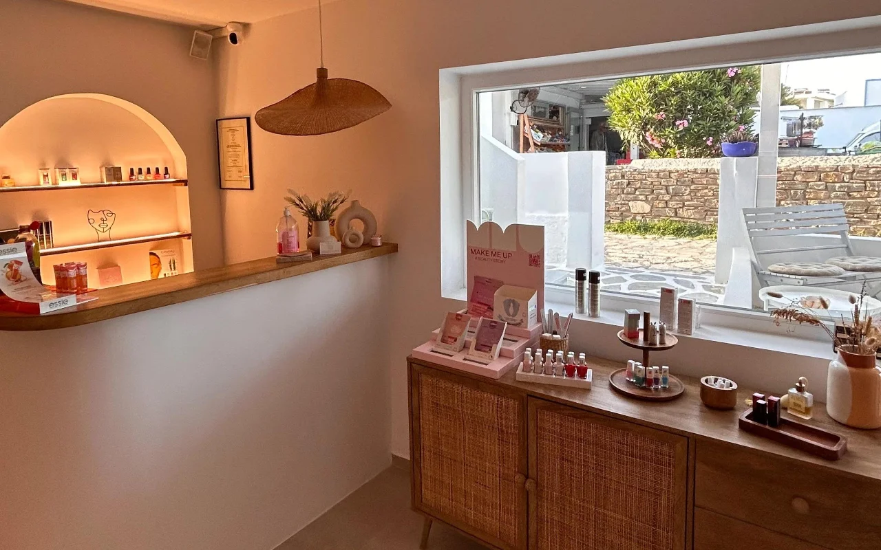 Το εσωτερικό του BeautiQ Esthetics and More στην Πάρο, με μια απαλά φωτισμένη τοξωτή κόγχη γεμάτη βερνίκια νυχιών, ένα πλεκτό κρεμαστό φωτιστικό ρατάν και ένα μεγάλο παράθυρο προς έναν πέτρινο τοίχο και έναν ηλιόλουστο κήπο.