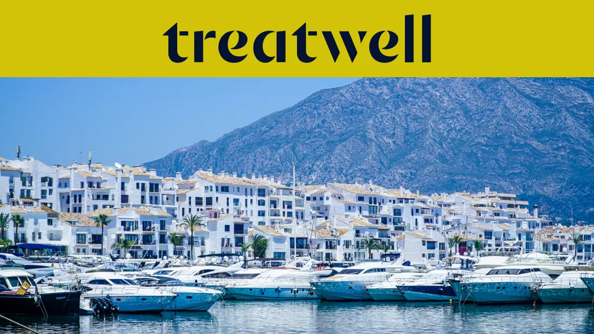 Η μαρίνα του Puerto Banús στη Marbella, με λευκά κτίρια διαμερισμάτων και αγκυροβολημένα σκάφη με φόντο τα βουνά, κάτω από ένα κίτρινο πανό Treatwell.