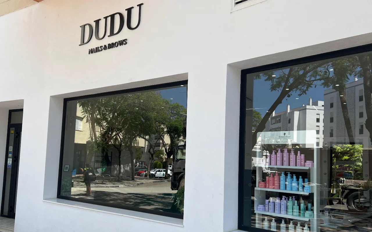 Η λευκή πρόσοψη του Dudu Nails and Brows στην Estepona, με το όνομα σε σκούρα γράμματα πάνω από φαρδιές βιτρίνες που δείχνουν σειρές προϊόντων μαλλιών.