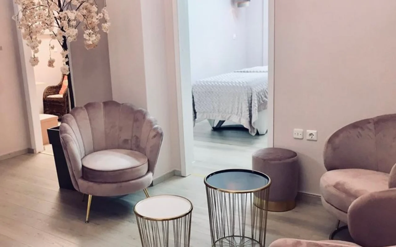 Ο απαλός ροζ χώρος αναμονής στο Nomikou Antonia Aesthetic Lab στη Σύρο, με μια βελούδινη πολυθρόνα με φεστόνια, λεπτά χρυσά βοηθητικά τραπεζάκια, ένα κλαδί ανθισμένης κερασιάς και ένα δωμάτιο περιποίησης πέρα από την πόρτα.