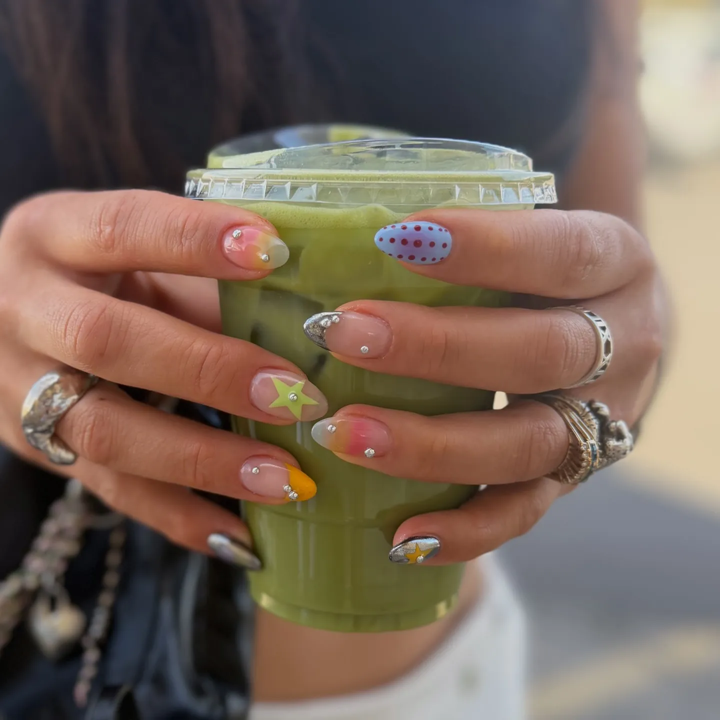 Ποικιλία από nail art με μικρά piercing από κρύσταλλο, μοτίβα αστεριού και πινελιές χρώματος σε νύχια σχήματος αμυγδάλου