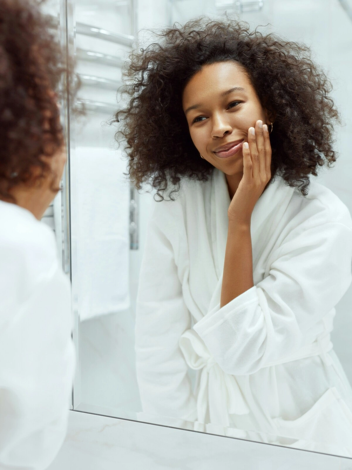 Μια γυναίκα με λευκό μπουρνούζι στέκεται μπροστά σε έναν μεγάλο καθρέφτη στο καθαρό, λευκό μπάνιο της και αγγίζει απαλά το λείο δέρμα του προσώπου της